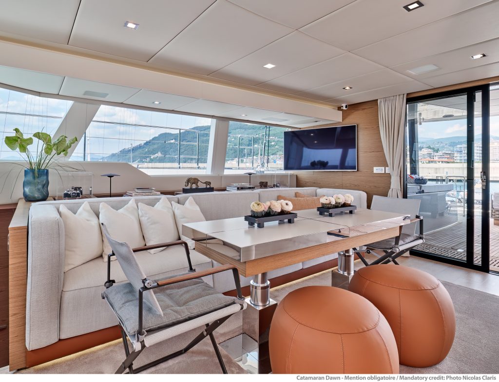 Widok wnętrza katamaranu Sunreef Yachts ukazujący luksusowy salon z jadalnią, dużym telewizorem i eleganckim wyposażeniem z widokiem na port przez duże okna.