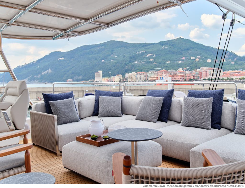 Luksusowy salon na katamaranie Sunreef Yachts z wygodnymi sofami i poduszkami, z widokiem na nadmorski krajobraz miejski z górą w tle.