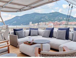 Luksusowy salon na katamaranie Sunreef Yachts z wygodnymi sofami i poduszkami, z widokiem na nadmorski krajobraz miejski z górą w tle.