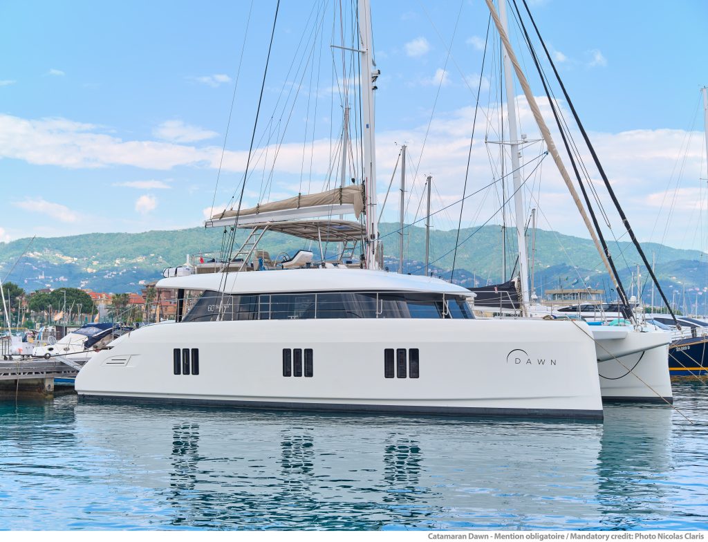 Luksusowy katamaran Sunreef Yachts o nazwie 'DAWN' zacumowany w marinie z innymi łodziami i górzystym tłem pod częściowo pochmurnym niebem.