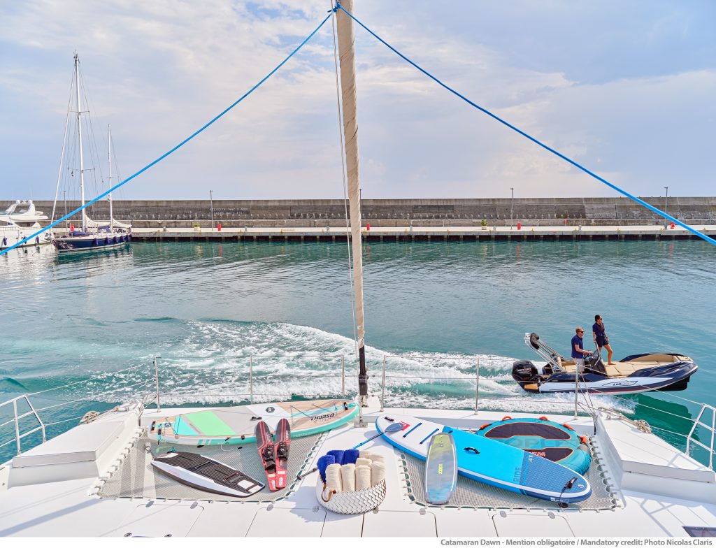 Widok z pokładu katamaranu Sunreef Yachts prezentujący jego luksusowe udogodnienia i krystalicznie niebieski ocean, z innym jachtem i długim molo w tle.