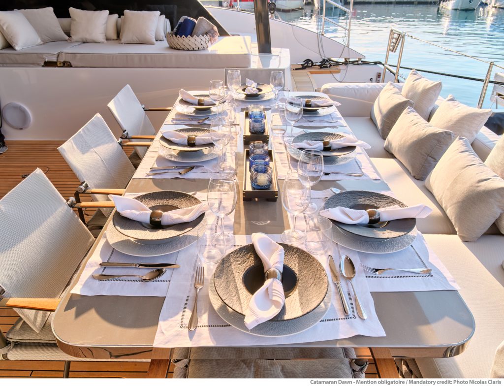 Elegancka aranżacja stołu na zewnątrz na katamaranie Sunreef Yachts, z pięknie nakrytym stołem i pluszowymi siedzeniami w tle.
