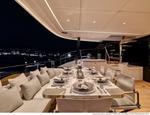 Luksusowa aranżacja jadalni na pokładzie katamarana Sunreef Yachts w nocy, z pięknie nakrytym stołem i wygodnymi miejscami do siedzenia, z widokiem na nadmorski krajobraz miejski.