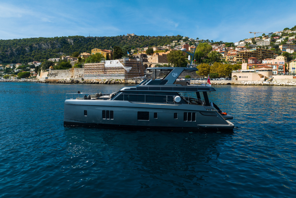 A luxurious Sunreef Yachts vessel is sailing near a coastal town with hills and numerous buildings in the background under a clear blue sky.