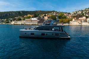 A luxurious Sunreef Yachts vessel is sailing near a coastal town with hills and numerous buildings in the background under a clear blue sky.