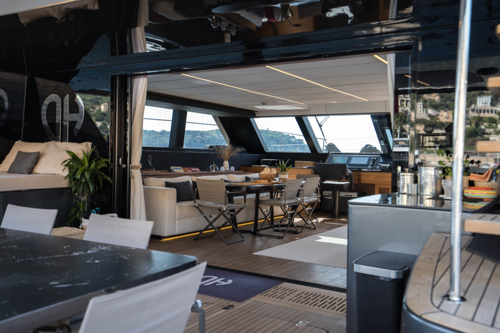 The interior view of a luxurious Sunreef Yachts catamaran. The spacious lounge features a dining table with chairs, a comfortable seating area, and a modern kitchen setup. Large windows provide a scenic view of the sea and coastline.
