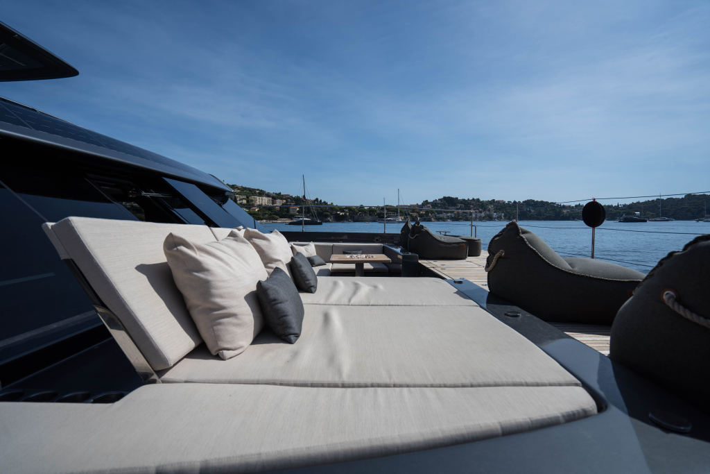Deck of a luxurious yacht with comfortable sunbeds and cushions, labeled as Sunreef Yachts, overlooking a calm sea and distant hilly landscape under a blue sky.