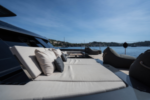 Deck of a luxurious yacht with comfortable sunbeds and cushions, labeled as Sunreef Yachts, overlooking a calm sea and distant hilly landscape under a blue sky.