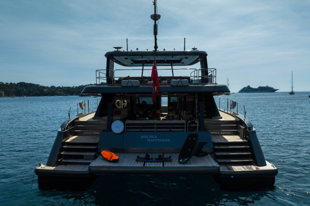 Back view of a luxury catamaran named 'Double Happiness' by Sunreef Yachts on the water, with a paddleboard and life preserver on deck.