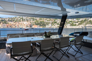 The image shows the outdoor seating area on a Sunreef Yachts catamaran. A large table with a white surface is surrounded by several chairs. The area overlooks a scenic harbor with sailboats and a hillside town in the background.