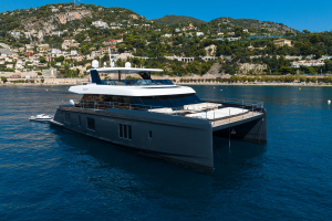 A large, luxurious catamaran yacht by Sunreef Yachts is anchored on calm blue waters. The yacht features multiple decks and modern design elements. In the background, there's a scenic coastline with lush greenery and hillside houses.