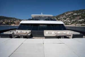 View of a luxury Sunreef Yachts catamaran's spacious deck with cushioned seating areas and wooden tables, set against a backdrop of coastal hills and a clear blue sky.