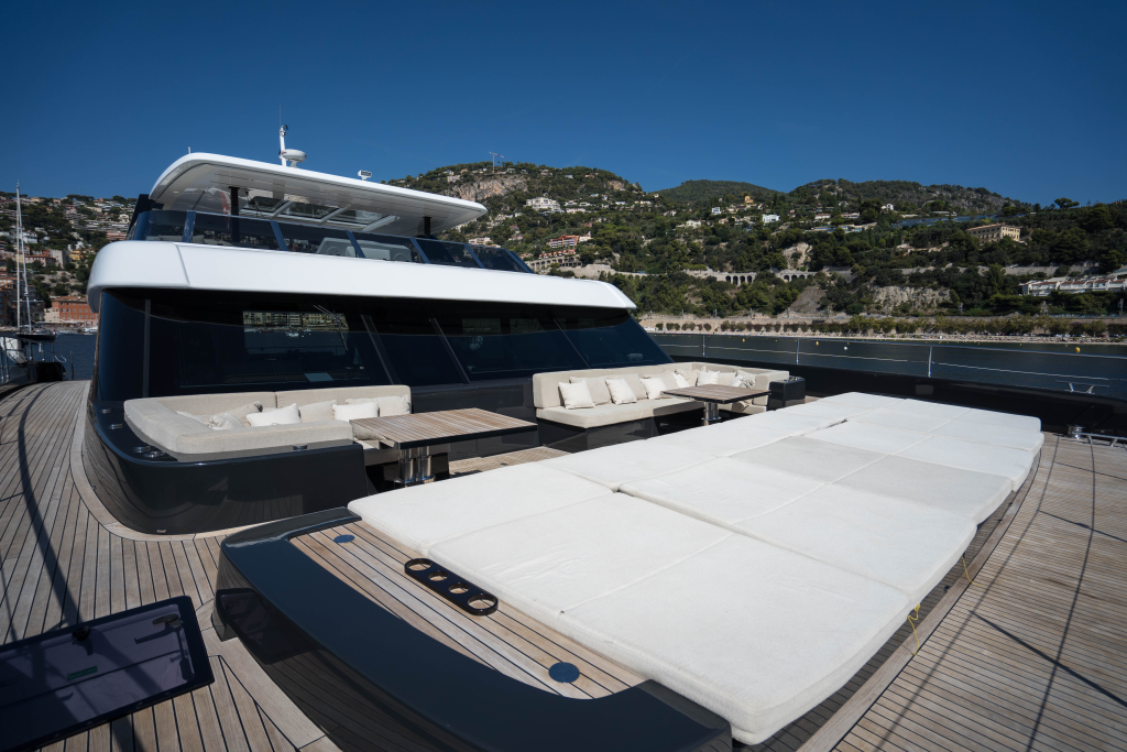 The image shows the spacious deck of a luxury Sunreef Yachts catamaran. The deck features large sunbathing loungers and comfortable seating areas, with hills in the background.