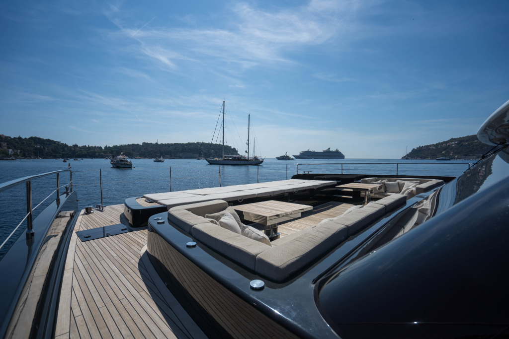 The image shows a luxurious yacht deck by Sunreef Yachts. The deck features comfortable seating areas with cream-colored cushions and a wooden table. Sailboats and a large cruise ship are anchored in the background under a clear blue sky.
