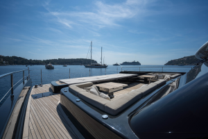 The image shows a luxurious yacht deck by Sunreef Yachts. The deck features comfortable seating areas with cream-colored cushions and a wooden table. Sailboats and a large cruise ship are anchored in the background under a clear blue sky.