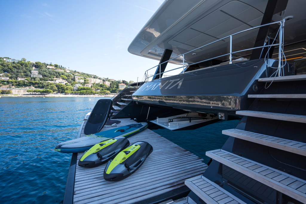 The image shows a luxurious Sunreef Yachts catamaran docked by a coastal area. The deck features modern amenities and there are surfboards on the platform. In the background, there's a view of the coast with green hills and buildings.