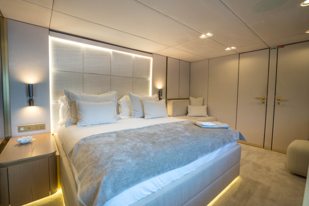 A luxurious bedroom interior on a Sunreef Yachts vessel, featuring a large bed with plush pillows and a soft beige and white color scheme. The room has subtle lighting and modern furnishings.