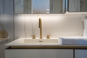 The image shows an elegant bathroom onboard a Sunreef Yachts vessel. It features a modern white sink with sleek gold fixtures, a countertop with neatly folded towels, and a beautifully lit mirror.