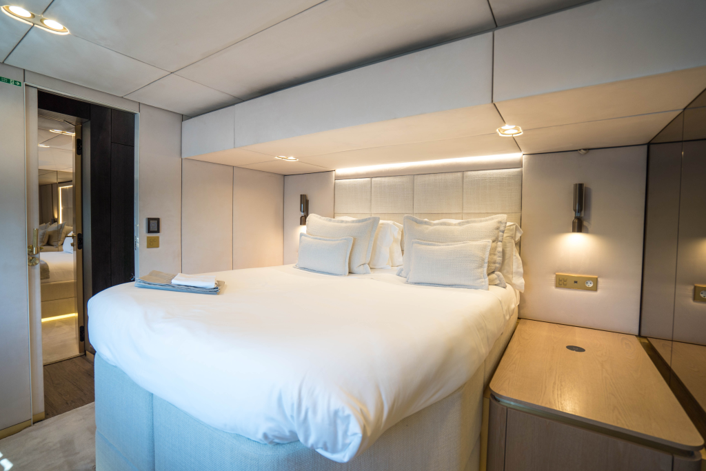 The image shows a luxurious cabin interior from Sunreef Yachts, featuring a large bed with crisp white linens and plush pillows. The space is well-lit with modern fixtures, and there's a wooden side table beside the bed.