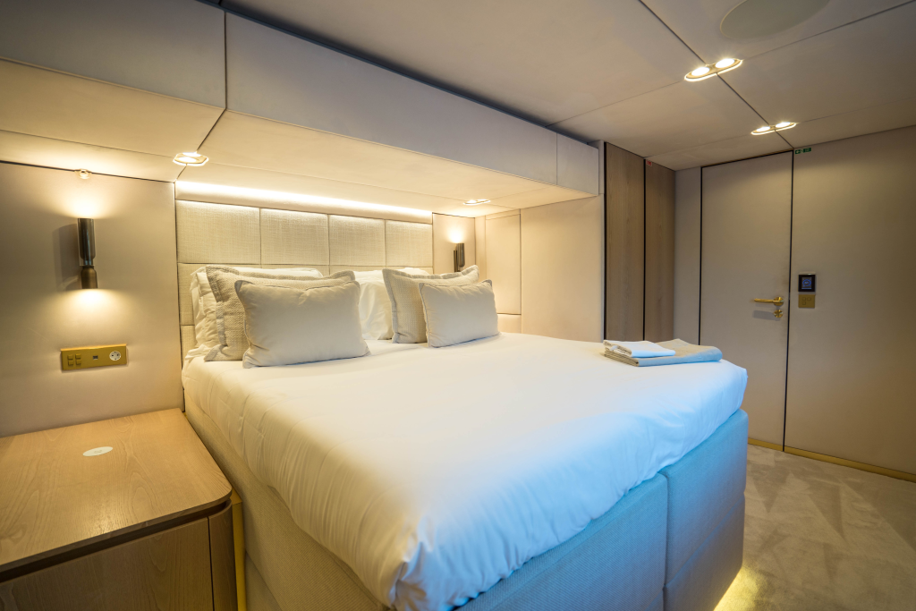 A luxurious bedroom inside a Sunreef Yachts vessel, featuring a plush double bed with white linens and soft pillows, illuminated by modern wall-mounted lights. The room showcases a sleek, minimalist design with light-colored wood accents and a small bedside table.