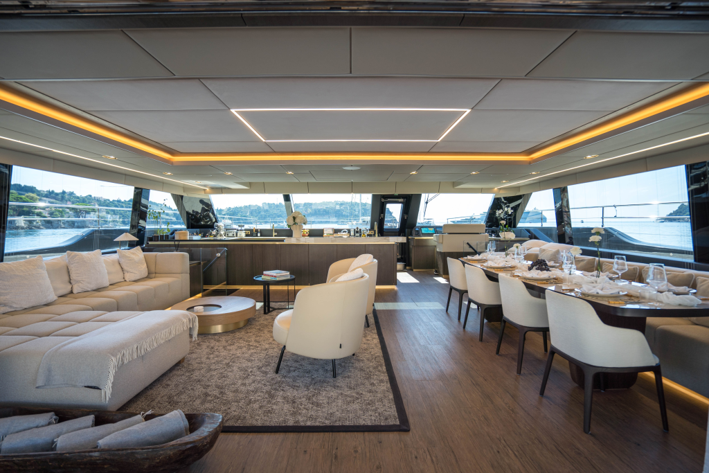 Interior of a luxurious Sunreef Yachts catamaran featuring a spacious salon with modern decor, a large seating area on the left, a dining table set for meals on the right, and wide windows offering a view of the sea.