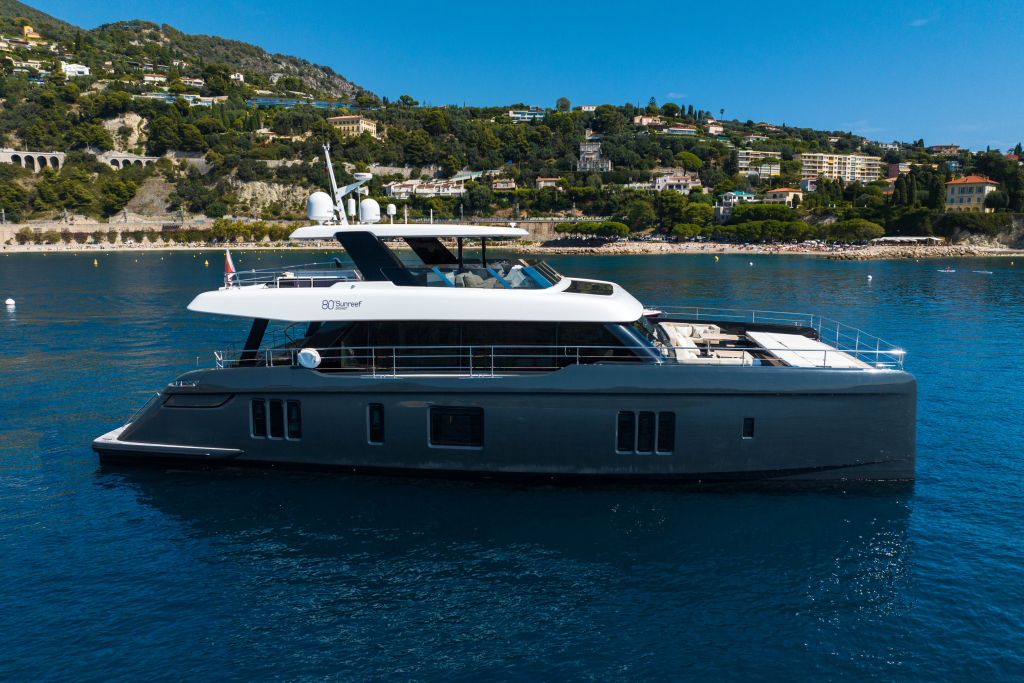 A luxurious Sunreef Yachts catamaran on clear blue water, with a scenic background of green hills and elegant villas.