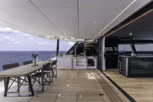 The image depicts the deck of a luxurious Sunreef Yachts vessel. It features an elegant, spacious seating area with a large wooden table and chairs, adjacent to a modern outdoor kitchen. The setting overlooks a calm sea under a partly cloudy sky.