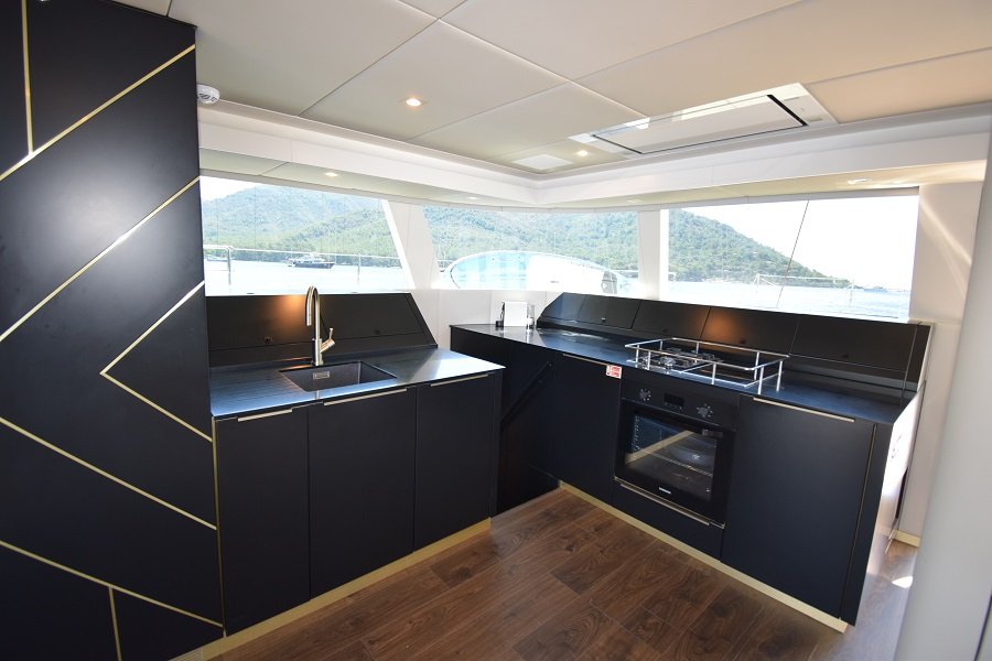 Obraz przedstawia nowoczesną kuchnię na katamaranie Sunreef Yachts. Zawiera eleganckie czarne szafki z złotymi akcentami, ciemny blat, wbudowany piekarnik i duże okna z widokiem na otaczającą wodę i wzgórza.