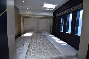 Wnętrze kajuty w Sunreef Yachts z podwójnym łóżkiem i wzorzystą narzutą, świetlikiem oraz dużymi oknami z widokiem na morze i góry.