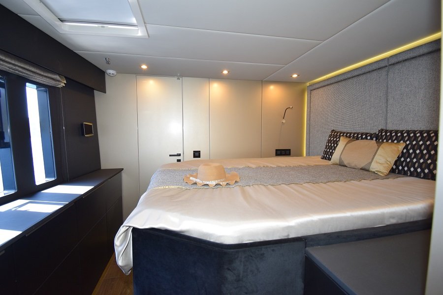 Wnętrze luksusowej kabiny jachtu Sunreef Yachts, z dużym łóżkiem z elegancką pościelą i kapeluszem na nim. Pomieszczenie posiada nowoczesne wyposażenie i nastrojowe oświetlenie.