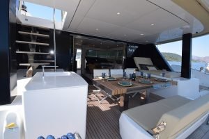 Obrazek przedstawia pokład luksusowego jachtu Sunreef Yachts. Na zewnątrz znajduje się drewniany stół z nakryciami i kieliszkami, otoczony wyściełanymi siedziskami. Schody prowadzą na górny pokład, w tle widoczne jest morze i odległe wzgórza.