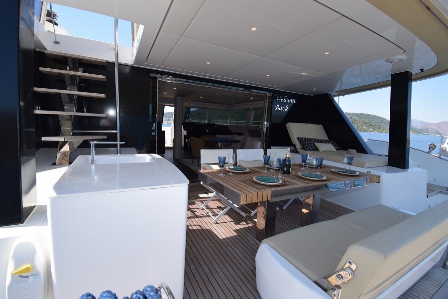 Obrazek przedstawia pokład luksusowego jachtu Sunreef Yachts. Na zewnątrz znajduje się drewniany stół z nakryciami i kieliszkami, otoczony wyściełanymi siedziskami. Schody prowadzą na górny pokład, w tle widoczne jest morze i odległe wzgórza.