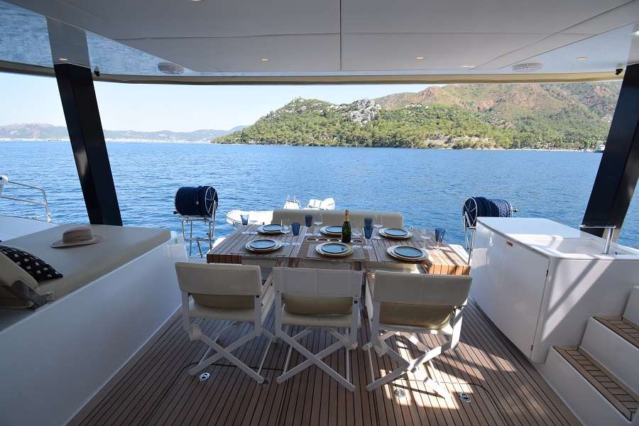 Zewnętrzny obszar jadalny na katamaranie Sunreef Yachts, z elegancko nakrytym stołem i krzesłami, z widokiem na spokojne niebieskie morze i odległe zielone wzgórza.