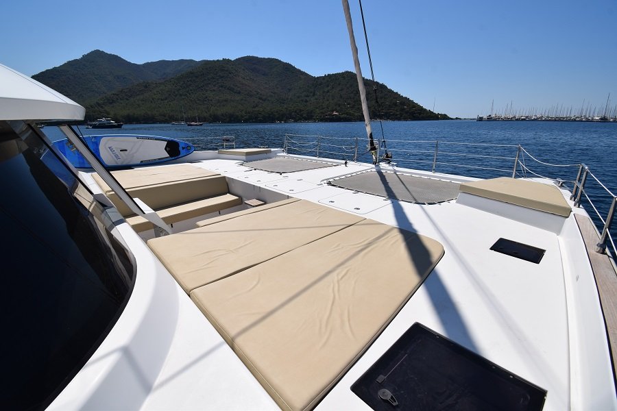 Pokład katamaranu Sunreef Yachts z beżowymi leżakami, otoczony spokojną, błękitną wodą i górami w tle.