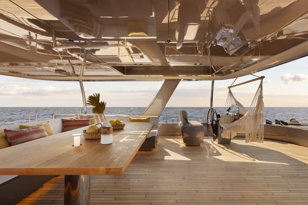 Obraz przedstawia luksusowy pokład Sunreef Yachts z eleganckim drewnianym stołem, dekoracyjnymi poduszkami i hamakiem. Pokład wychodzi na rozległy ocean pod czystym niebem.