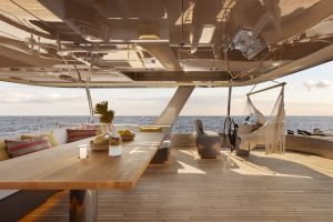 Obraz przedstawia luksusowy pokład Sunreef Yachts z eleganckim drewnianym stołem, dekoracyjnymi poduszkami i hamakiem. Pokład wychodzi na rozległy ocean pod czystym niebem.