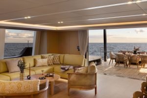 Wnętrze luksusowej łodzi Sunreef Yachts, z przestronną częścią wypoczynkową wyposażoną w komfortowe siedzenia i drewniany stolik kawowy. Okna od podłogi do sufitu oferują widok na otwarte morze i zewnętrzną jadalnię, podkreśloną eleganckimi meblami drewnianymi i delikatnym oświetleniem.