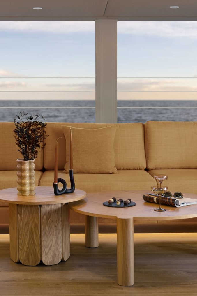 Eleganckie wnętrze salonu Sunreef Yachts z jasną drewnianą podłogą i meblami. Dwa okrągłe drewniane stoliki zdobią nowoczesne dekoracje, świece i mały wazon z dekoracyjnymi gałązkami. Duża beżowa kanapa stoi przed szerokimi oknami, z widokiem na ocean.