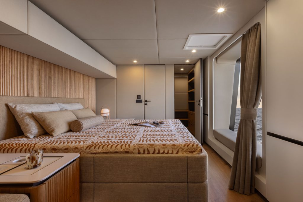 Luksusowo i elegancko zaprojektowane wnętrze sypialni na jachcie Sunreef Yachts, z dużym łóżkiem z wzorzystą pościelą, stylowymi panelami drewnianymi, nastrojowym oświetleniem i oknem z widokiem na ocean.