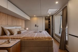 Luksusowo i elegancko zaprojektowane wnętrze sypialni na jachcie Sunreef Yachts, z dużym łóżkiem z wzorzystą pościelą, stylowymi panelami drewnianymi, nastrojowym oświetleniem i oknem z widokiem na ocean.