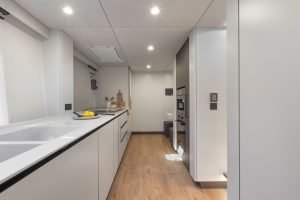 Wnętrze nowoczesnej kuchni na katamaranie Sunreef Yachts, z eleganckimi szafkami, urządzeniami ze stali nierdzewnej i drewnianą podłogą. Na blacie znajduje się miska z cytrynami.