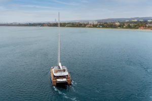A Sunreef Yachts catamaran sails in calm waters with a coastline and cityscape visible in the distance.