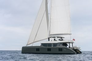 A modern catamaran with white sails navigates through calm waters under a cloudy sky. It has a sleek, dark-colored hull and a spacious upper deck. A small boat is attached at the stern.
