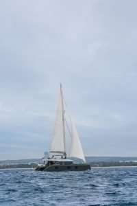 A luxury sailing catamaran by Sunreef Yachts is gliding over the water with its sails unfurled, set against a cloudy sky.