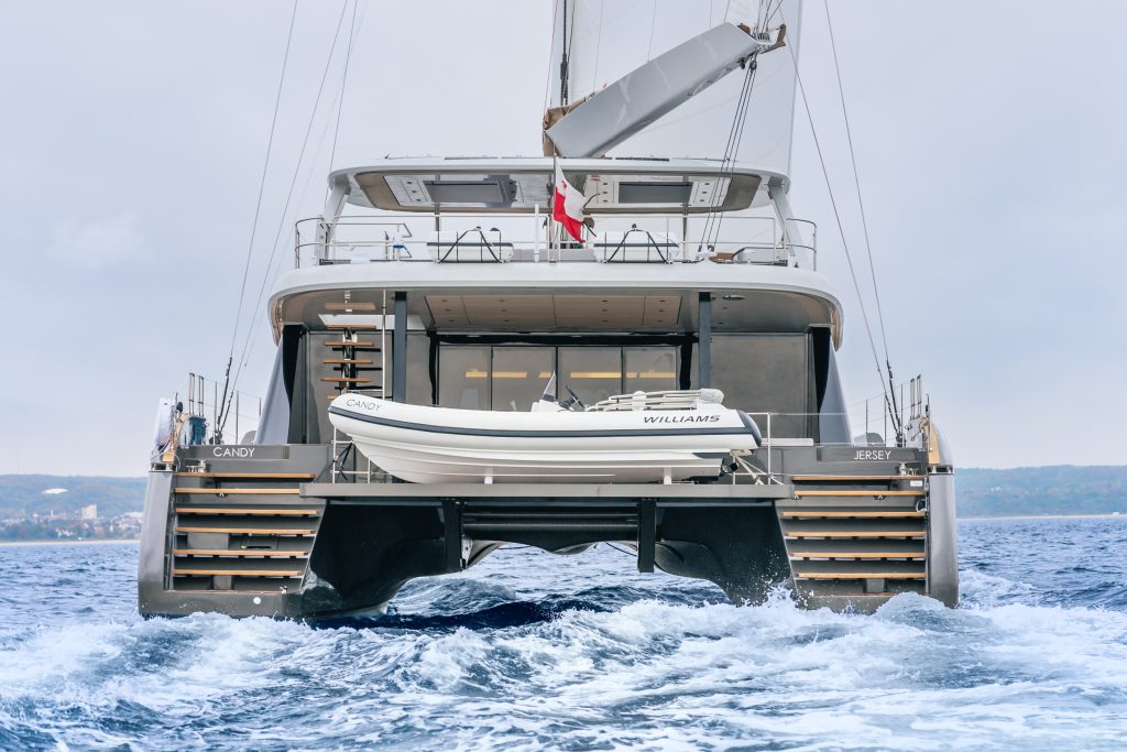 A Sunreef Yachts catamaran is sailing on the ocean, showing its rear design with an additional small boat named Williams. The name Candy is visible on the side.
