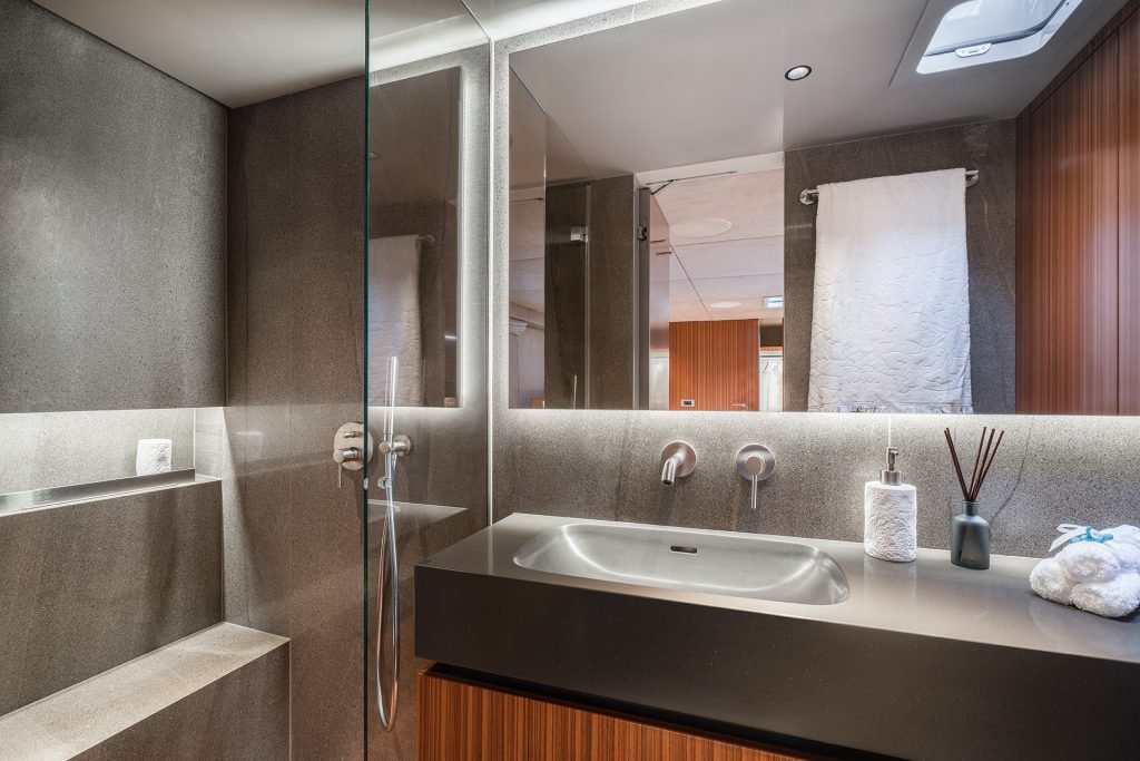 The image shows a modern bathroom on a Sunreef Yachts vessel. The space features sleek grey walls, a glass-enclosed shower, and a minimalist vanity with a metallic sink. A rolled towel and toiletries are neatly arranged nearby.