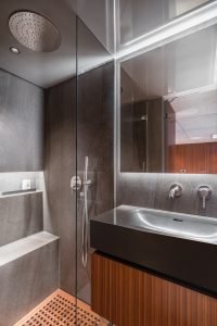 A modern bathroom on a Sunreef Yachts vessel features a sleek design with a large glass shower, a stainless steel overhead shower head, a dark stone sink, and wooden accents. The space is compact yet luxurious, emphasizing contemporary aesthetics.