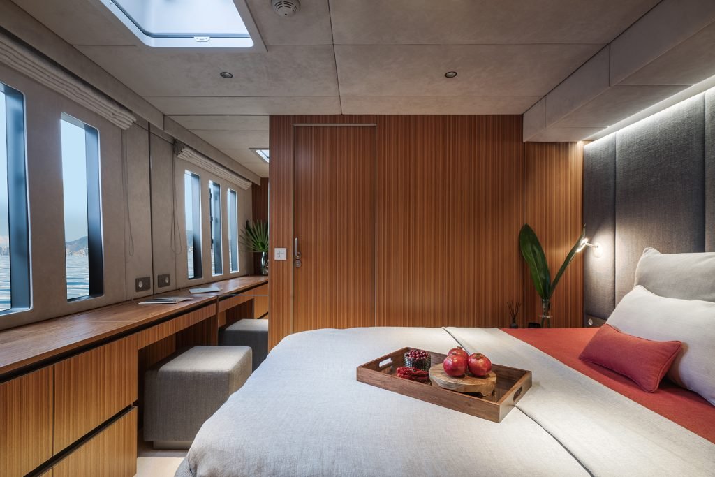 A luxurious bedroom on a Sunreef Yachts boat, featuring a large bed with white and red bedding. A wooden tray with fruit is on the bed. Windows on the left offer a view of the sea. The room has modern wooden paneling and elegant decor.