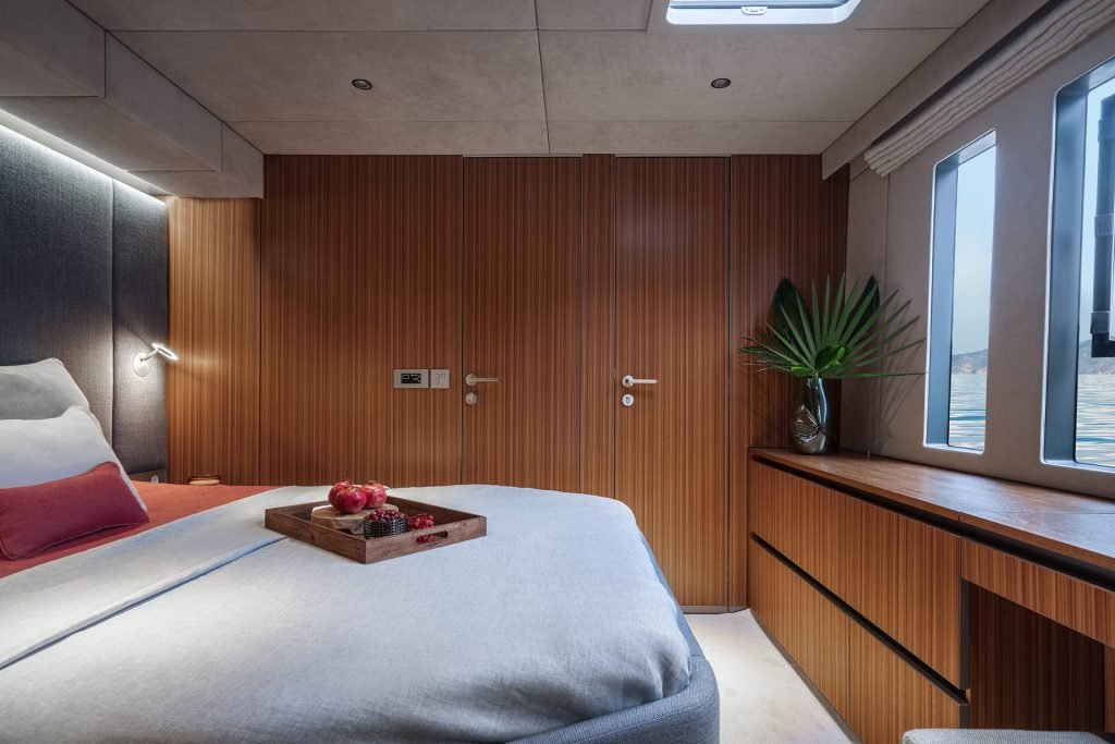 Interior of a Sunreef Yachts cabin featuring a cozy bed with white linens and a wooden tray holding fruit. The walls and cabinetry are made of rich wood, and a window provides a view of the water.