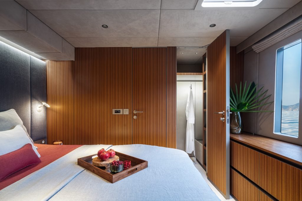 A luxurious bedroom interior on a Sunreef Yachts vessel features a cozy bed with a tray holding pomegranates and blackberries. The room has wooden paneling, a small cabinet with a plant in a vase, and a window showing a view of the sea.